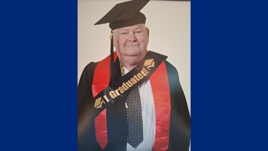 Ray is wearing a graduation gown and hat, and a sash that says I graduated