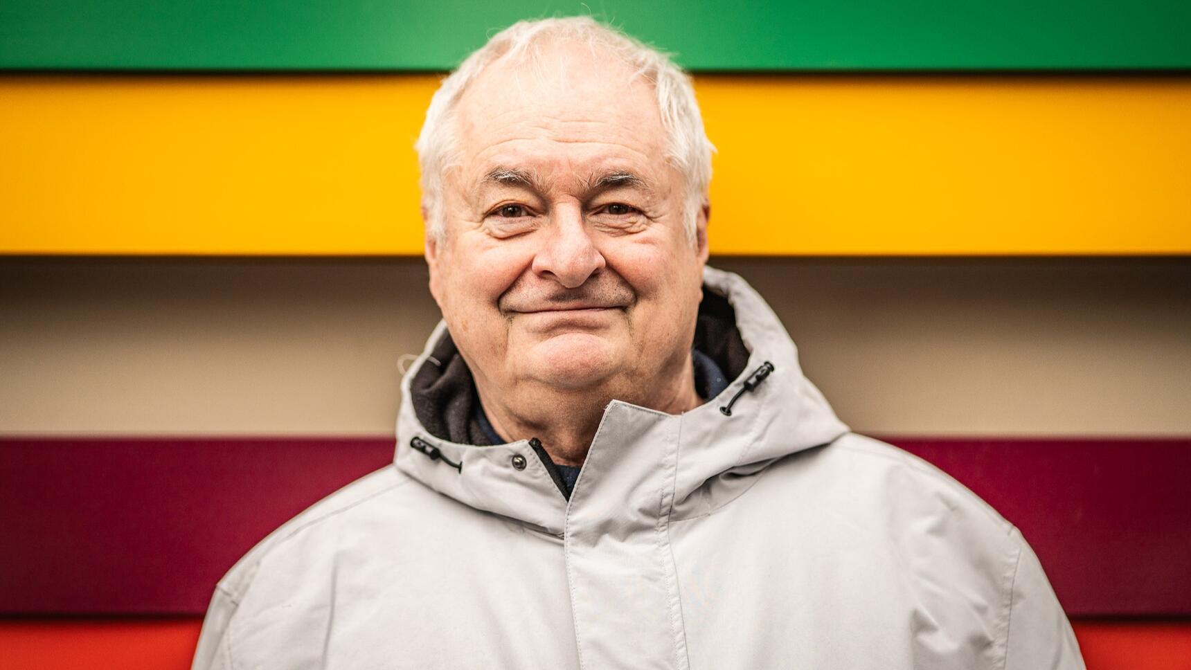 A man in front of coloured stripes wearing a grey raincoat