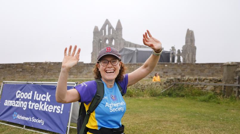 Woman waving as she walks part Whitby Abbey ruins.