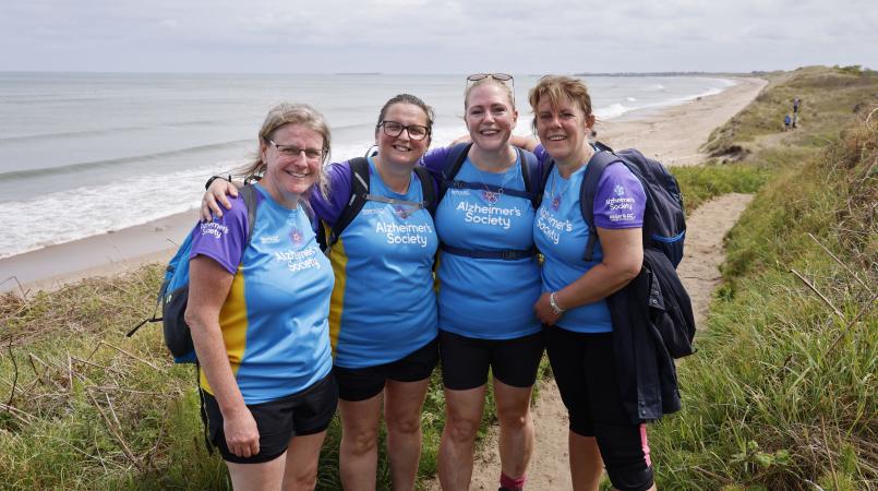 Four women stood on a coastal path hugging and smiling.