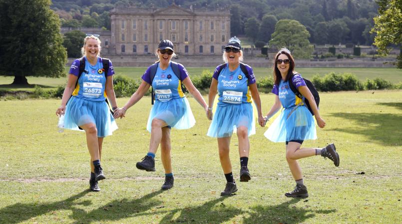 Four women in blue tutus dancing on a grassy field.