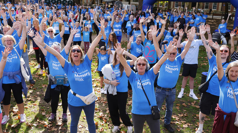 A crowd of walkers in blue Memory Walk t-shirts cheer with their hands in the air in front of the inflatable start line at Bristol Memory Walk