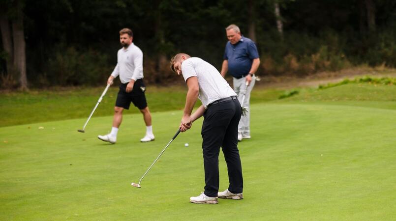 Three men playing on a golf course, one takes a golf shot