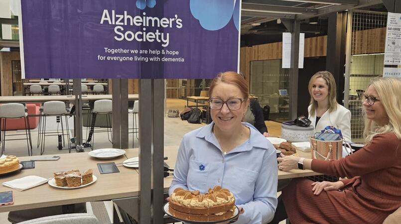 A woman with ginger hair, a blue shirt and glasses holding a cake and smiling