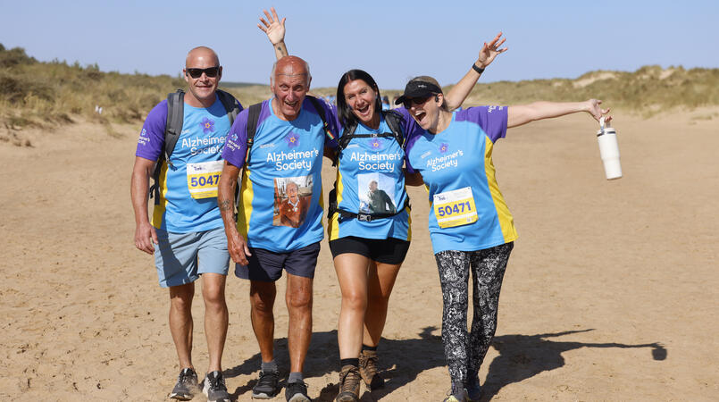 A group of 4 smiling at the camera as they walk along a beach.