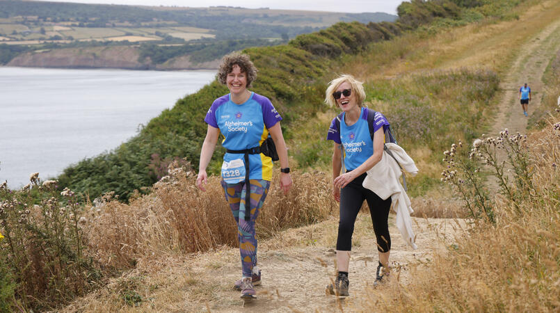 2 people smiling at the camera as they walk up a hill next to the sea.