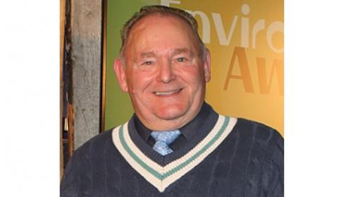 A smiling Ernie looking at the camera wearing a blue tie under a blue jumper