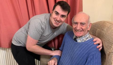 Tom smiling beside his Grandad who sits in an armchair