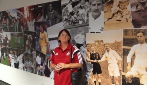 Sue wearing a red football shirt standing in front of a wall of large football photos