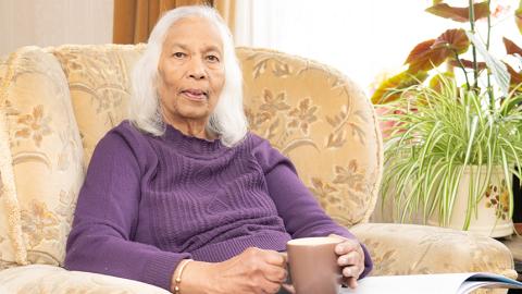 Shirley wears a purple jumper while sat in her armchair holding a mug and smiling