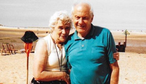 Eileen is cuddled close to Peter while standing on a beach as the tide is out