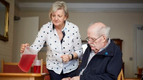 A woman pouring juice out of a jug for a man with dementia sat at at a table