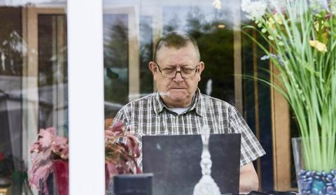 An older man sits at the window in his living room, reading something on his laptop