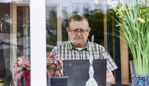 An older man sits at the window in his living room, reading something on his laptop