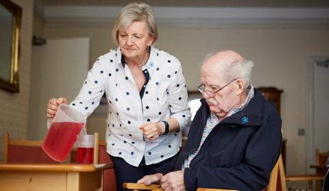 Elderly person with dementia seated next to a carer who is pouring them a drink from a jug into a cup