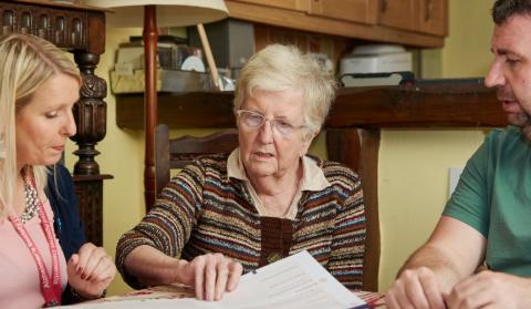 A professional and family member sit either side of an older woman, talking her through some paperwork