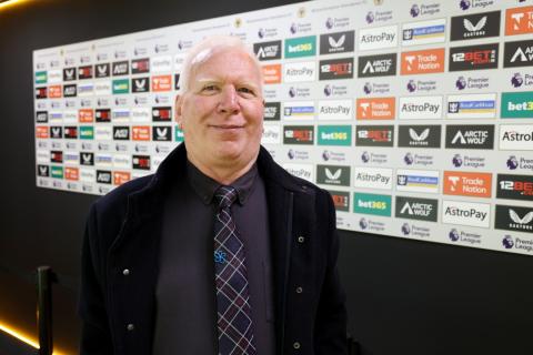 Peter Jones stood in front of a wall with advertisements on it at a football stadium, wearing an Alzheimer's Society pin badge