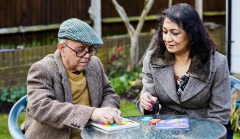 An older man and younger woman sit in the garden reading and playing a game