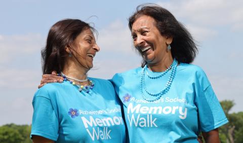 Two women wearing Alzheimer's Society Memory Walk t shirts are laughing. One woman has her arm round the other