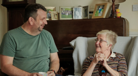 A woman in an armchair looks up at her adult son who is sitting next to her.