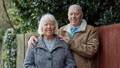 A man and woman stand smiling outside in winter coats