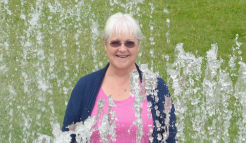 A woman in sunglasses stands amongst fountains of water.