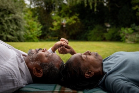A woman and man lie on their backs on the grass, holding hands.