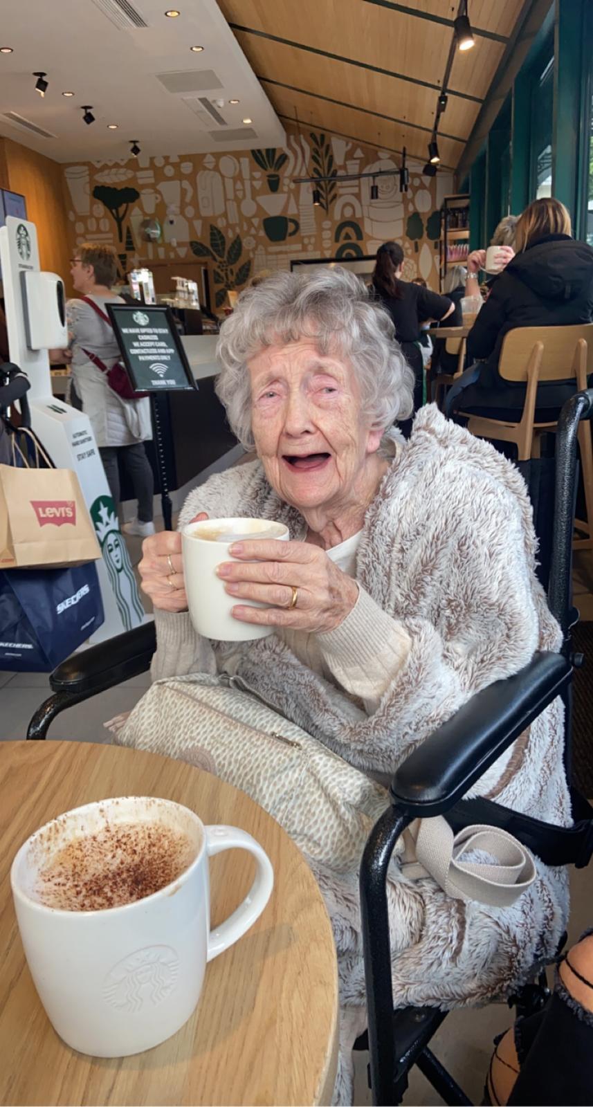 A woman with short grey hair wearing a grey top drinking a warm drink in a cafe