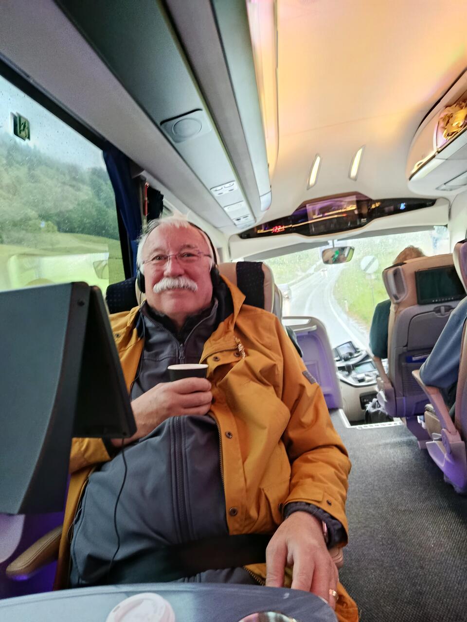 A man with white hair and moustache wearing a yellow coat and drinking a coffee on a coach