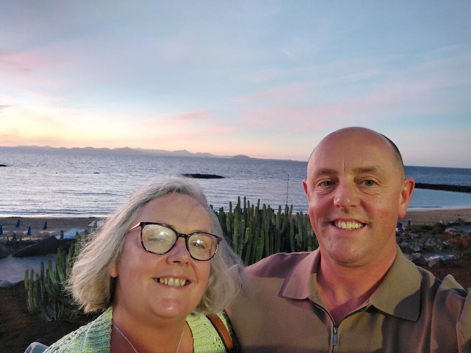A woman with short grey hair and glasses and a bald man with a green top on a beach
