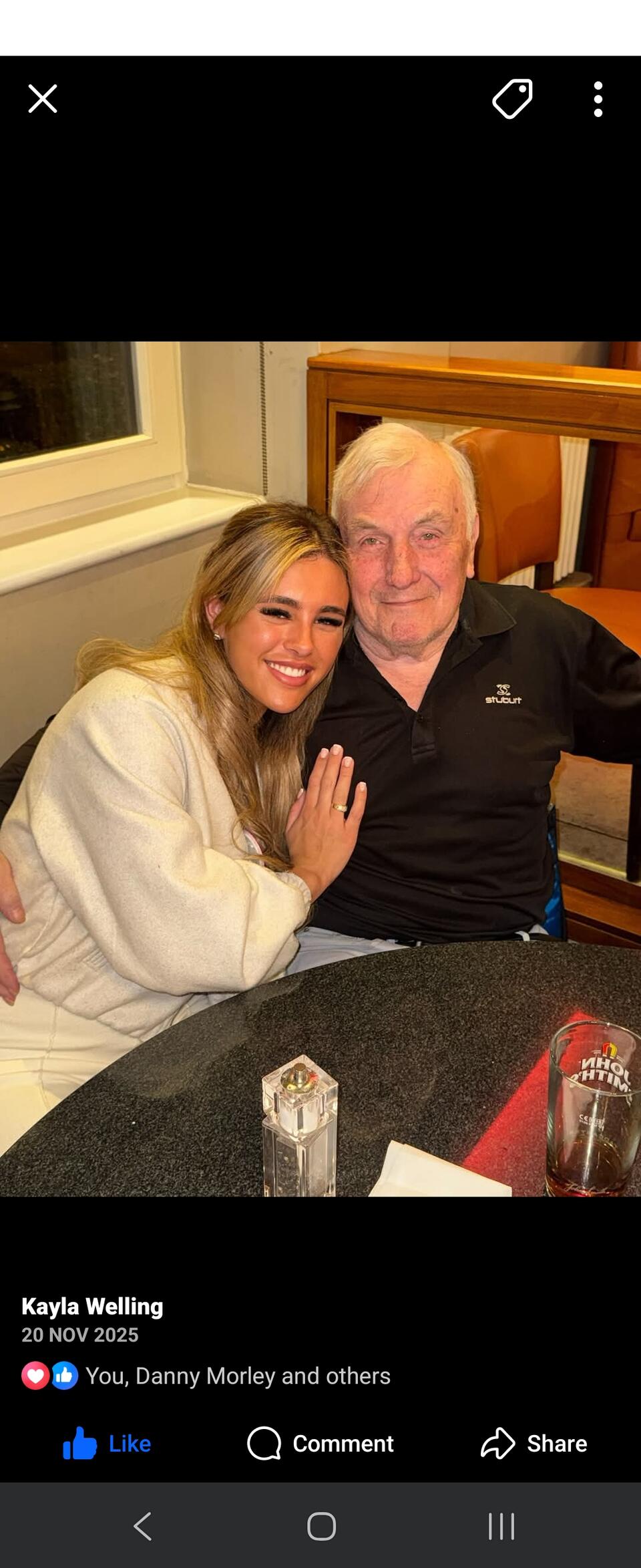 A younger woman with long blonde hair wearing a white jumper hugging an older man with white hair wearing a blue t shirt
