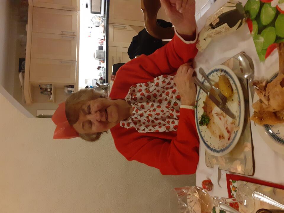 A woman wearing a red patterned top, red cardigan and Christmas cracker hat eating a roast dinner