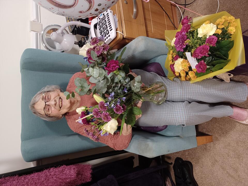 A woman with short grey hair, pink top and grey trousers sat in a green chair holding flowers