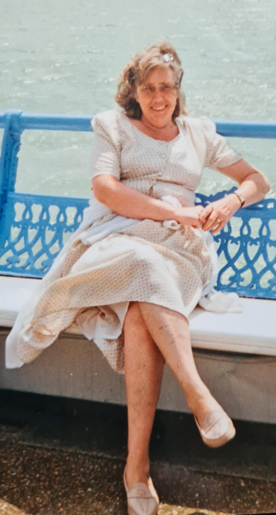 A woman with light hair wearing a cream dress sat on a bench by the sea