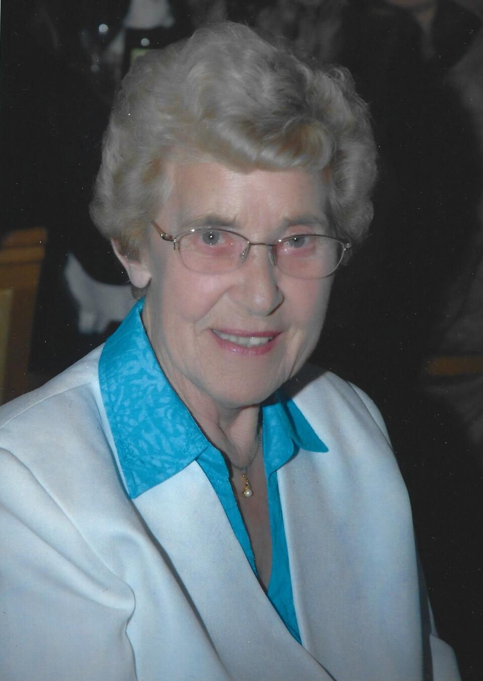 A smiling woman with short, grey hair, wearing glasses and blue shirt and white blazer
