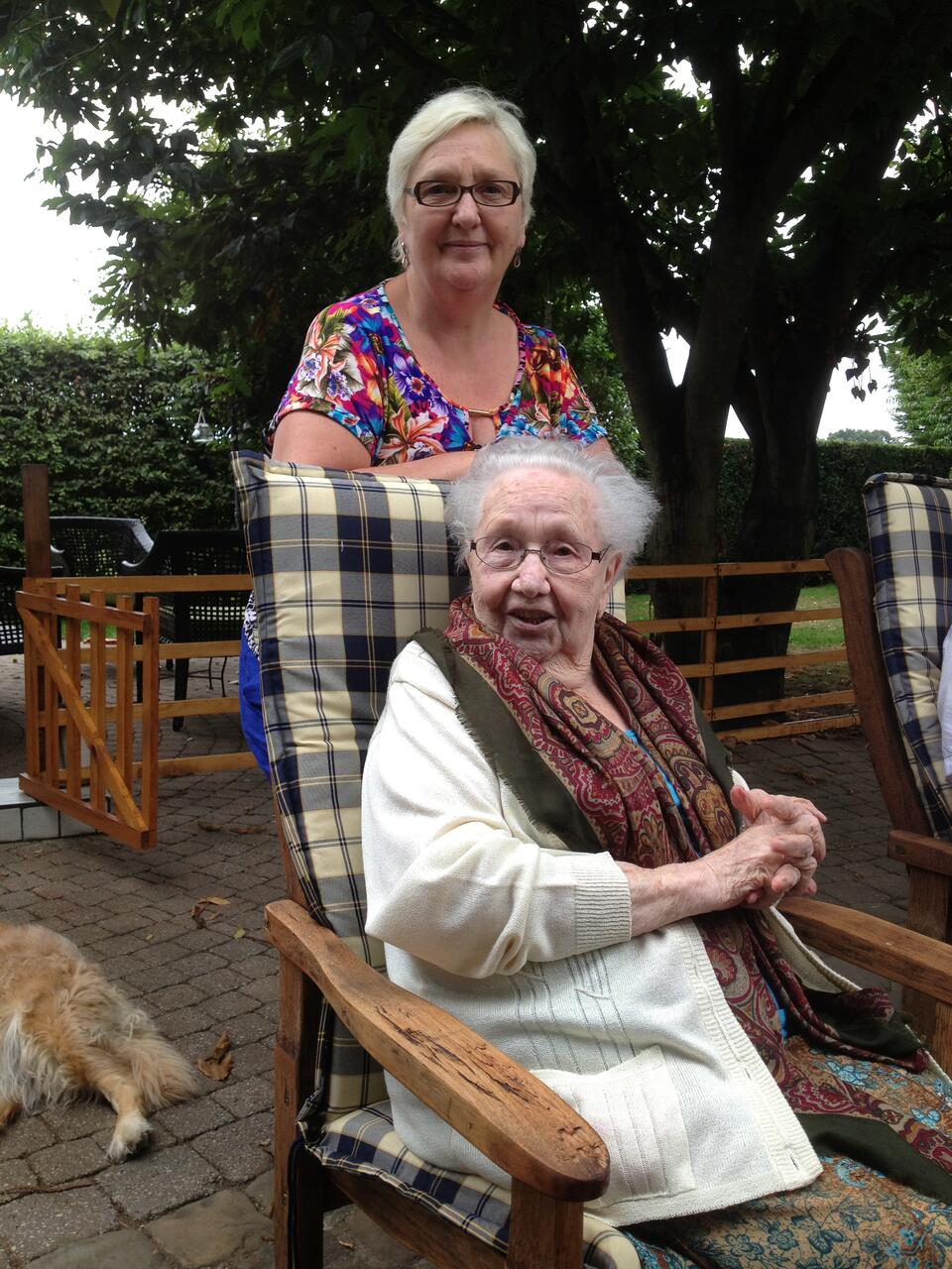 An older woman sat on a chair in a garden with a younger woman stood behind her