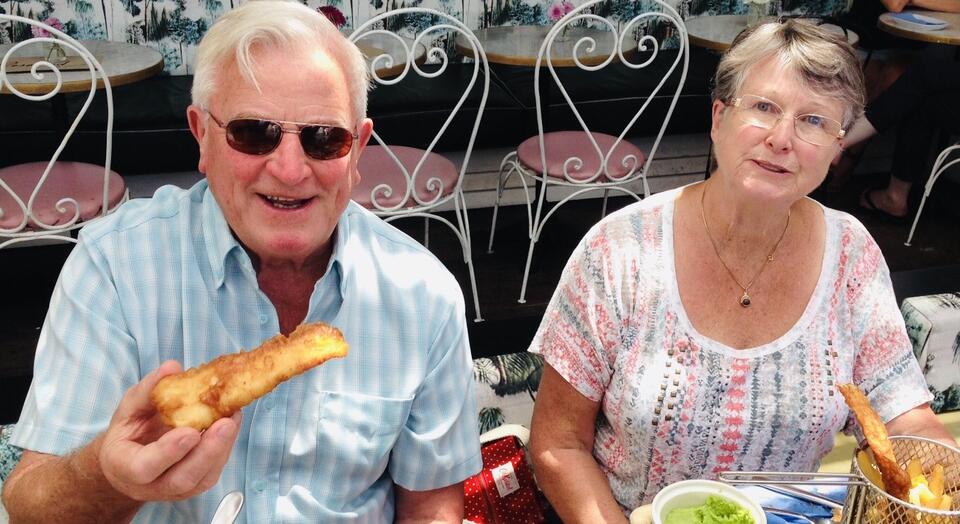 A man with white hair, sunglasses and a light blue shirt holding battered fish and a woman with white and grey hair and patterned top