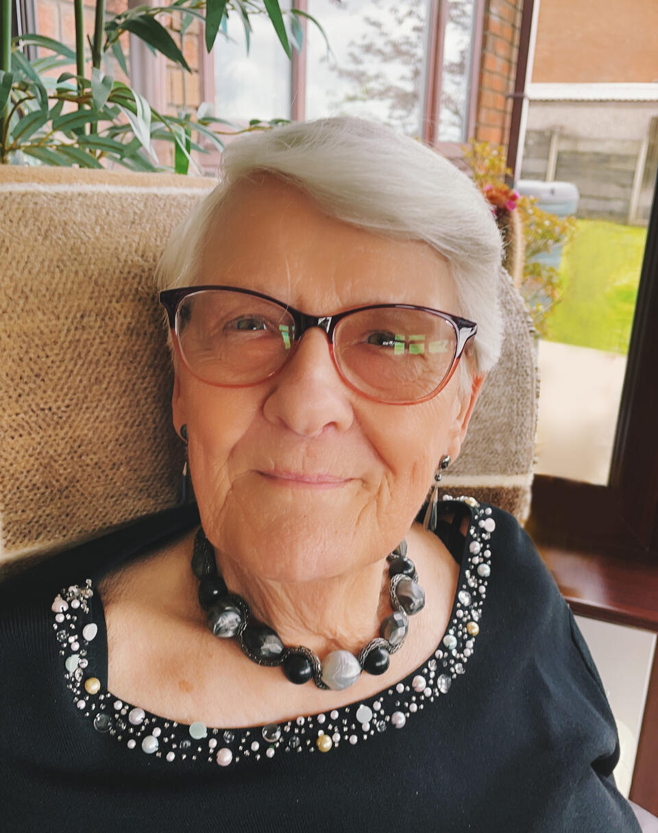 A woman with short grey hair, black glasses, black necklace and black top sat in a chair