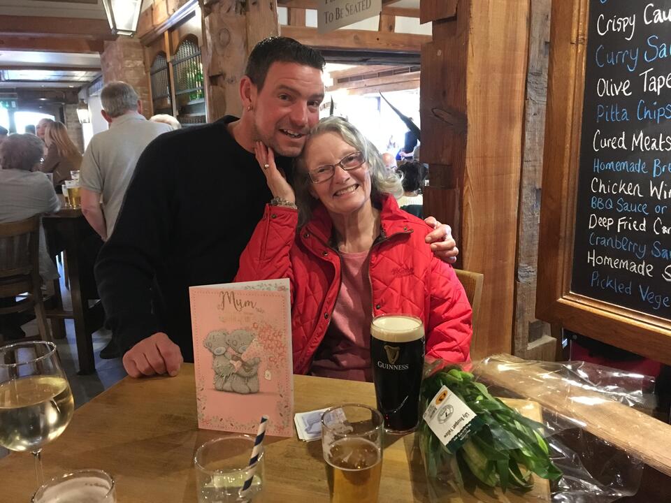 A man with brown hair and dark top and a woman with light wavy hair wearing a red coat in a pub