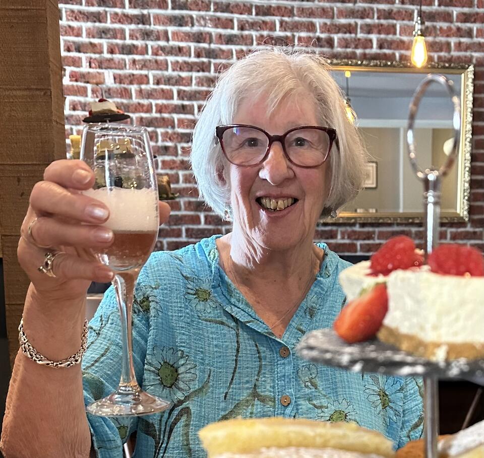 A woman with short white hair, glasses and a blue shirt holding a glass of prosecco