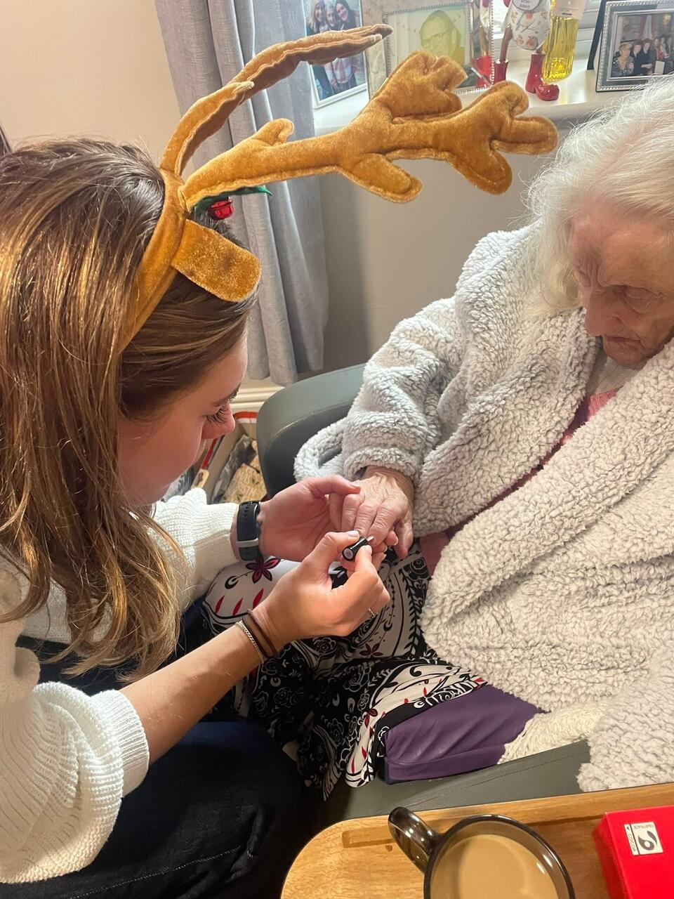 A woman wearing reindeer antlers painting the nails of an older woman sat in a chair