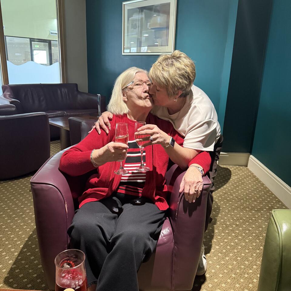 A woman behind a chair kissing a woman sat in the chair