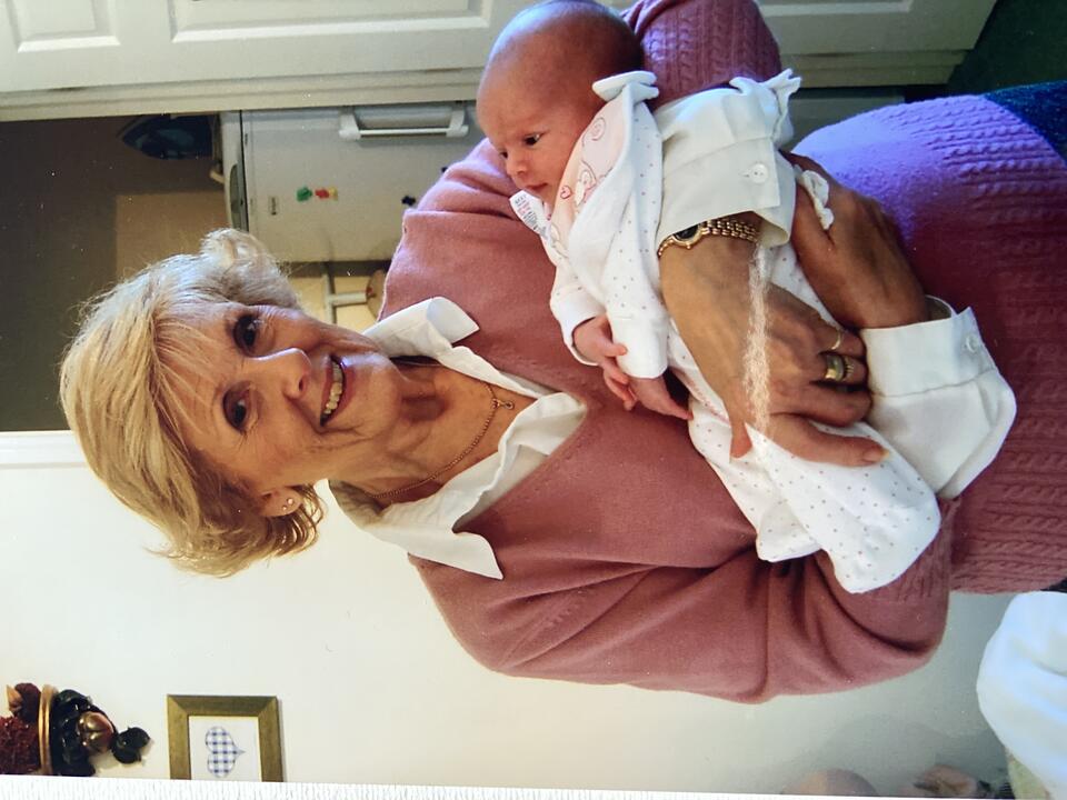A woman with short blonde hair wearing a white shirt and pink jumper holding a baby dressed in white