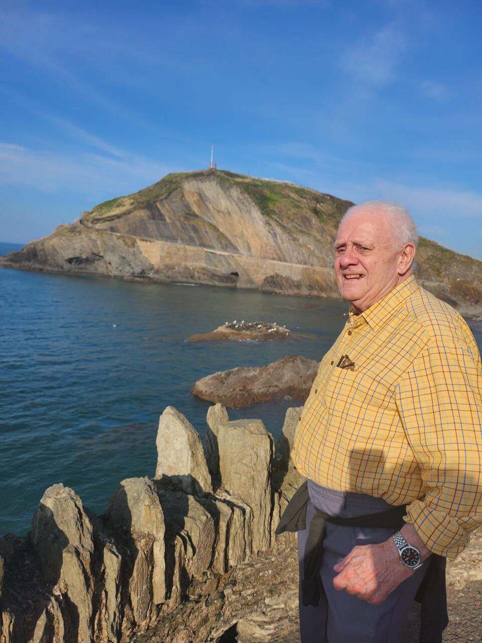 A man wearing a yellow shirt stood in front of sea and a hill