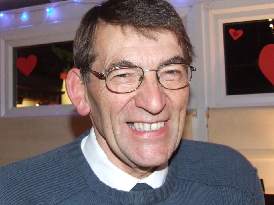 A smiling man with brown hair wearing glasses, a white shirt and blue jumper