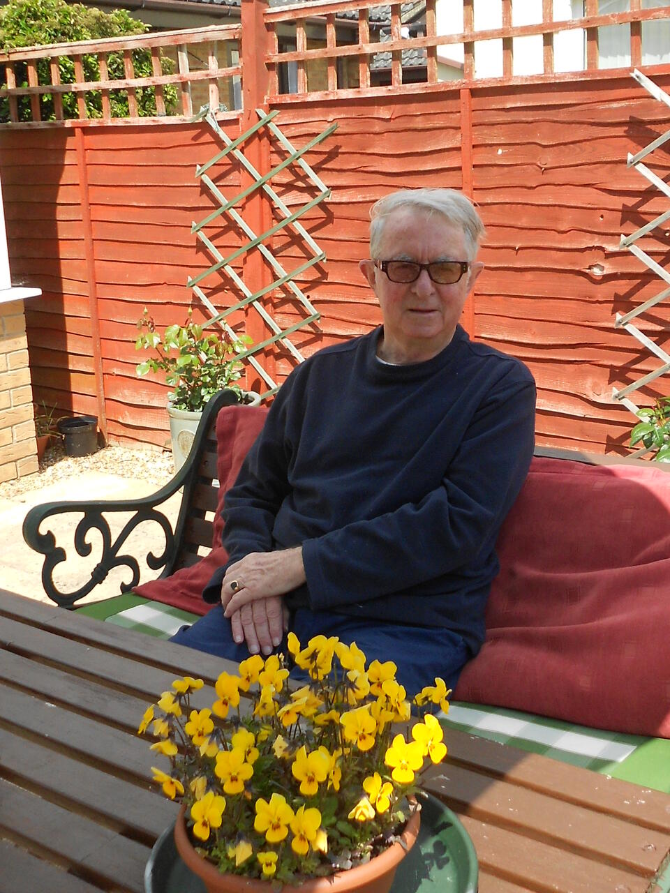 A man with white hair, sunglasses and dark jumper sat on a bench in a garden