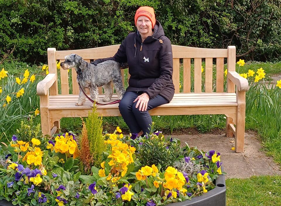A woman with an orange hat, brown coat and jeans sat on a bench with a small dog