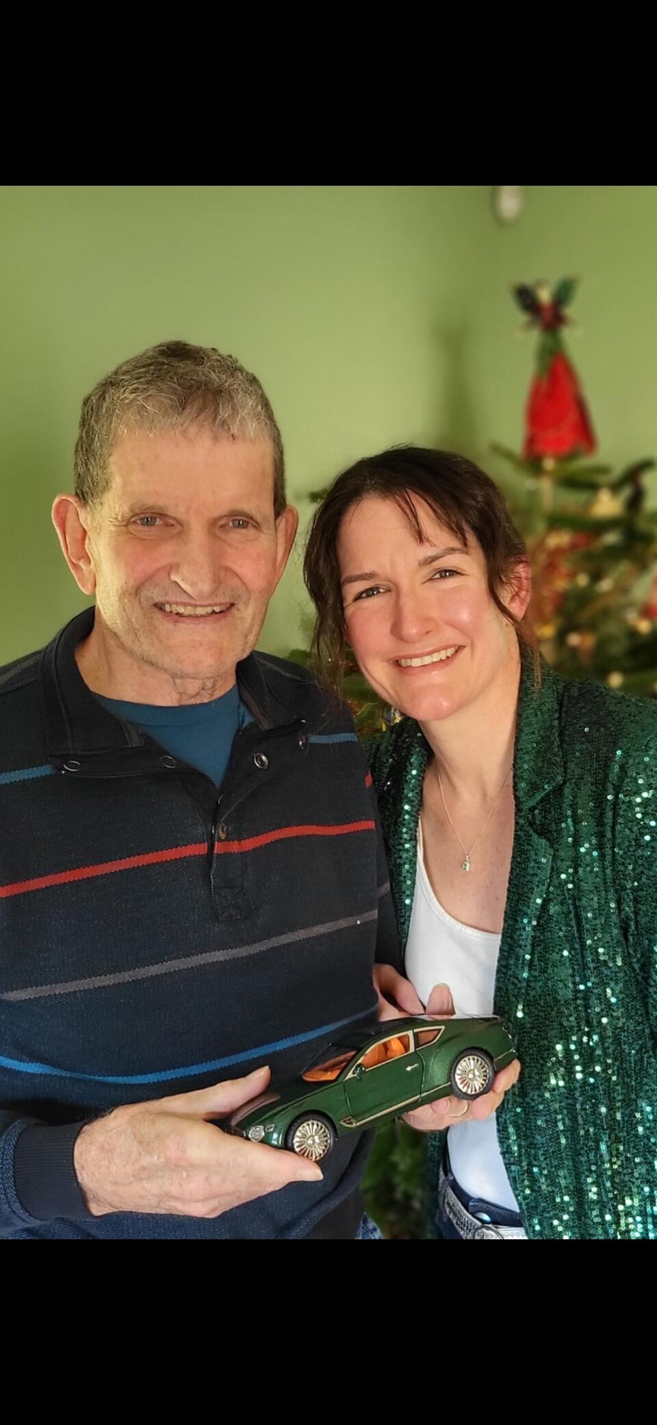 A man, holding a model car, and woman smiling, a Christmas tree is in the background