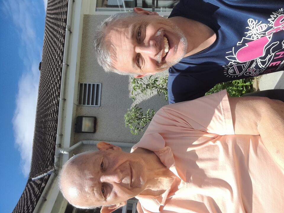 A bald man wearing a peach shirt and a man with grey hair wearing a blue patterned t-shirt in a garden