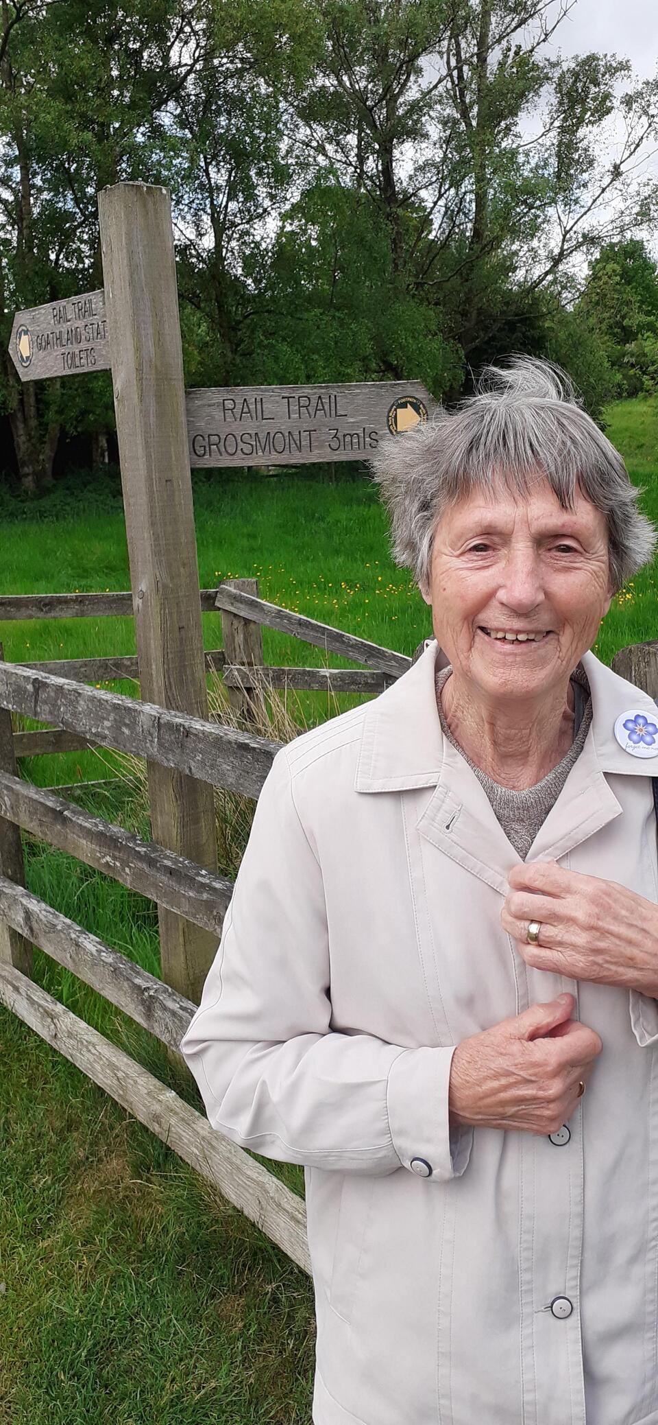 A lady with grey hair smiling. She's wearing a white coat and is stood in front of a sign
